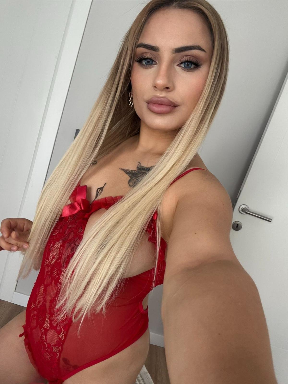 Self-portrait of a woman with long blonde hair wearing red lace lingerie, standing in a doorway.