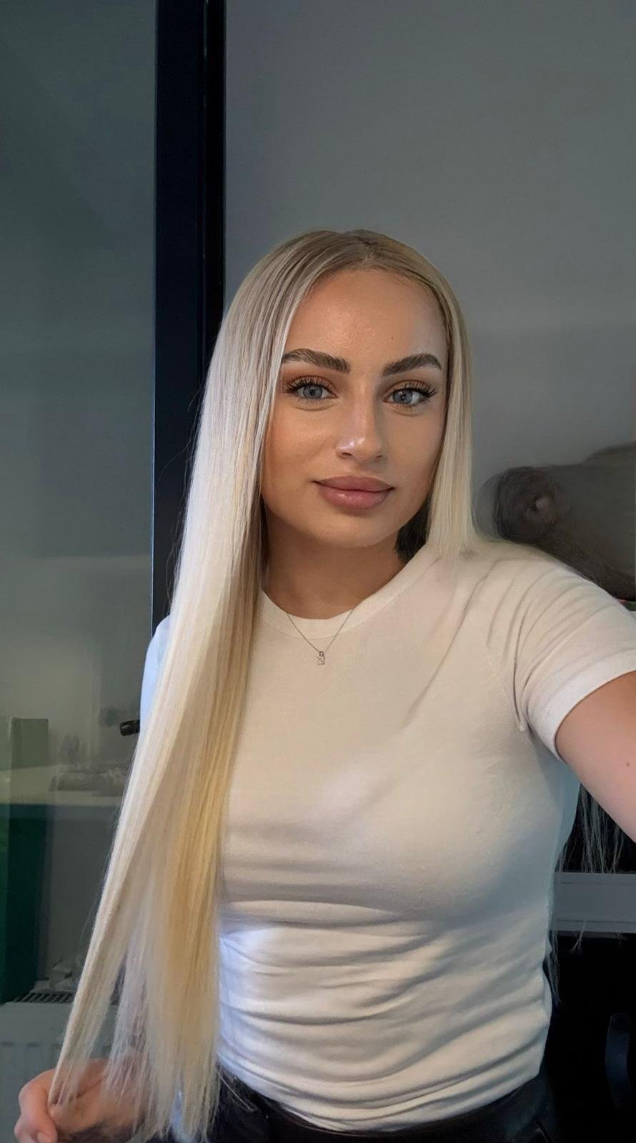 Selfie of a young woman with long platinum-blonde hair and a white t‑shirt, smiling softly at the camera indoors.