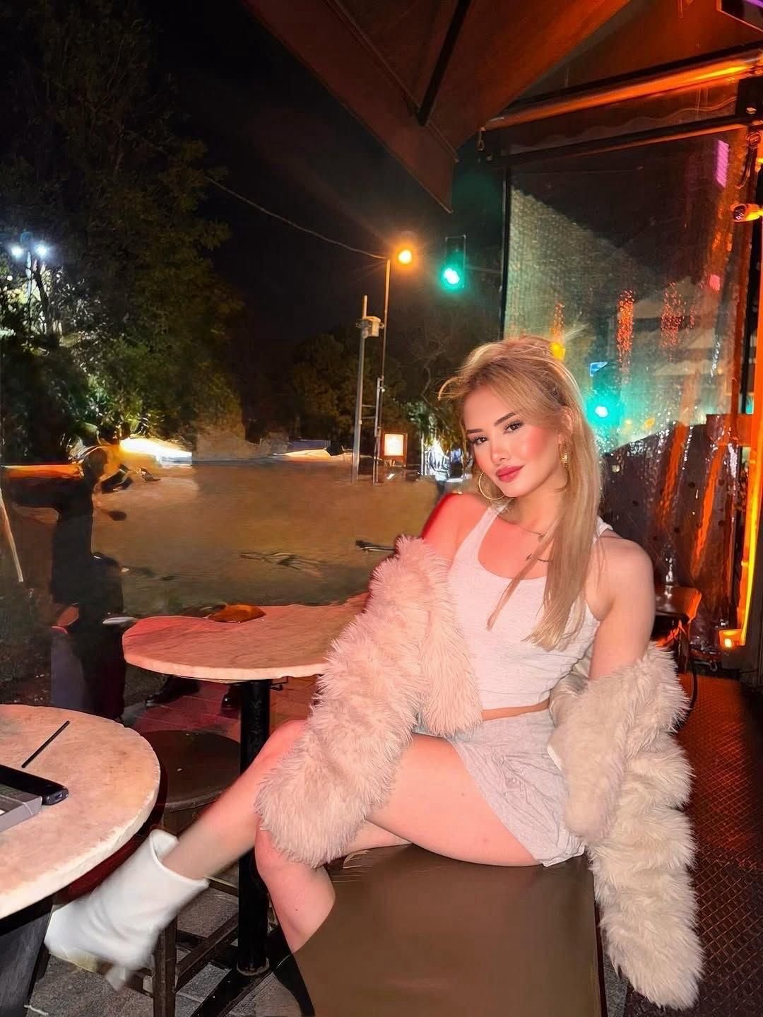 Woman in white crop top and fluffy coat sits at an outdoor cafe table at night, posing with city lights in the background.