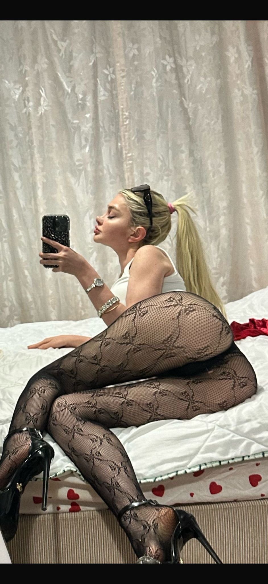 Blonde woman taking a mirror selfie on a bed, wearing a white top, black floral fishnet tights and high heels.