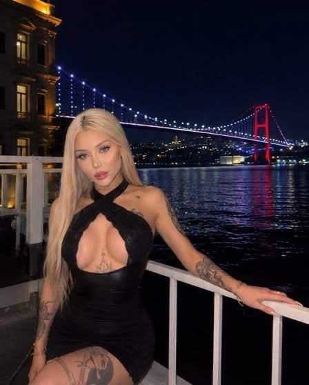 Blonde woman in a black cut‑out dress posing on a railing at night, with a lit suspension bridge and water in the background.