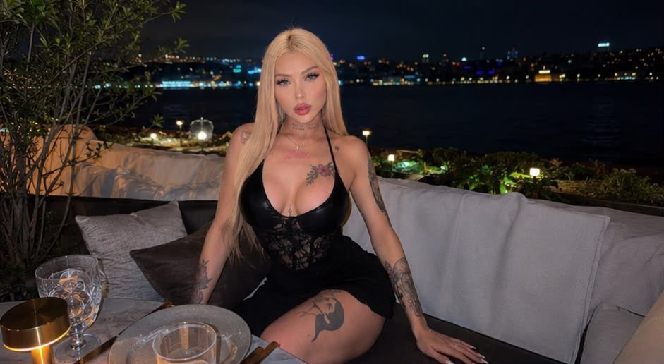 Blonde woman in a black lace outfit sits at an outdoor waterfront restaurant with city lights in the background at night.
