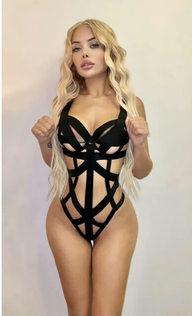 Woman with long blonde hair wears a black geometric cut-out bodysuit and poses with fists raised near chest.