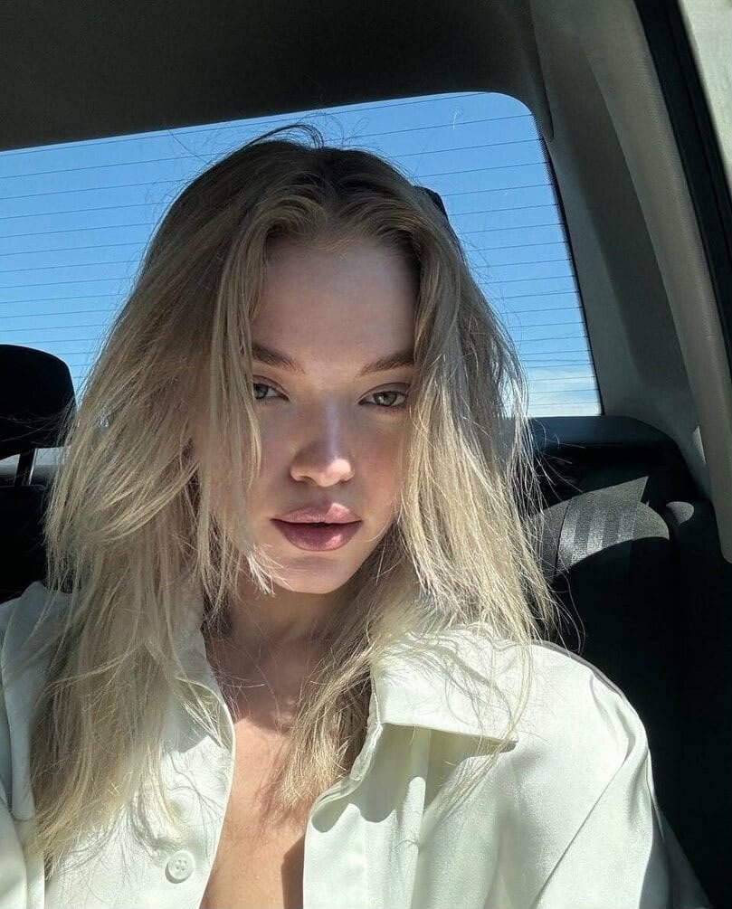 Close-up of a woman with blonde hair in a light shirt, sitting in a car with sunlight on her face.