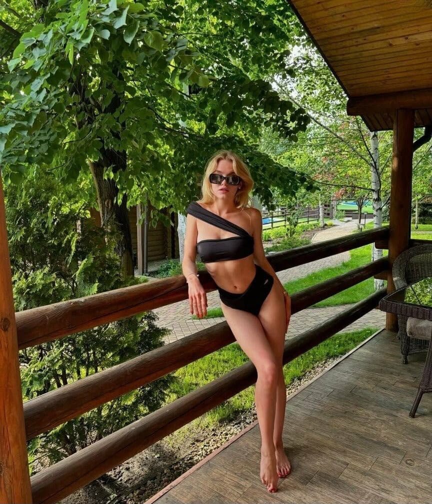 Woman in a black two-piece swimsuit with sunglasses leaning on a wooden railing of a covered porch, greenery in the background.