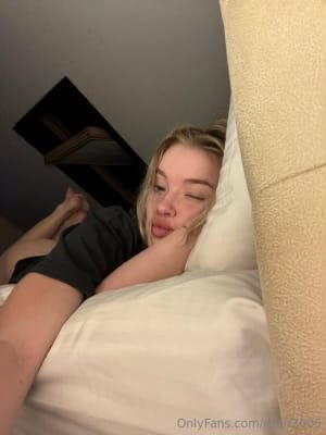 Young woman lying on a bed with a white pillow, resting her head to the side and puckering her lips toward the camera.