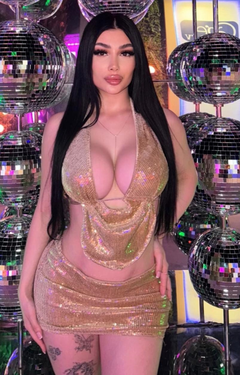 Woman with long black hair wearing a gold sequin cut‑out dress posing among disco balls in a club.