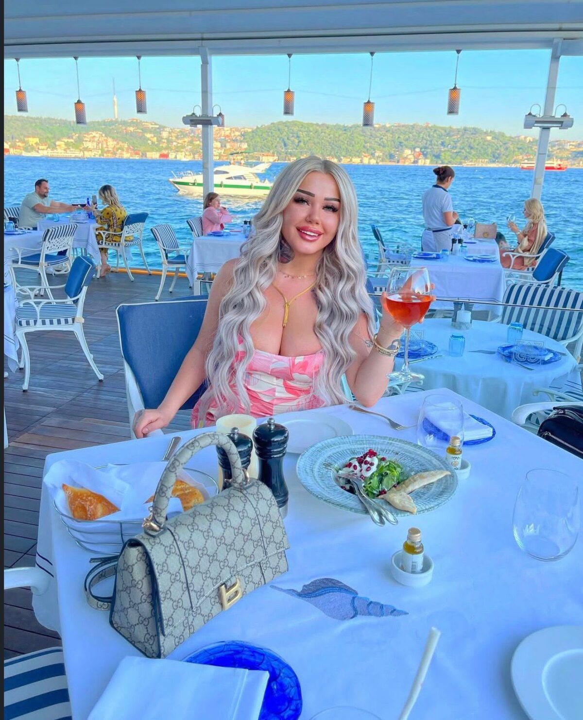 Woman at an outdoor seaside restaurant on a boat, holding a glass of rosé and smiling, with plates and a Gucci handbag on the table against blue water and hills in the background.