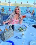 Woman with long blonde hair in a pink dress sits at a seaside restaurant aboard a boat, smiling while holding a glass of rosé; a Gucci handbag and plated food are in the foreground with blue water and hills in the background.