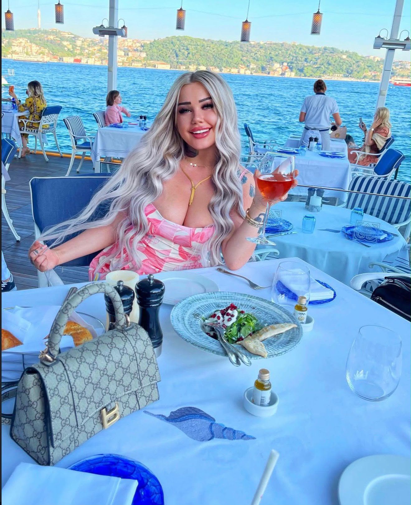 Woman with long blonde hair in a pink dress sits at a seaside restaurant aboard a boat, smiling while holding a glass of rosé; a Gucci handbag and plated food are in the foreground with blue water and hills in the background.