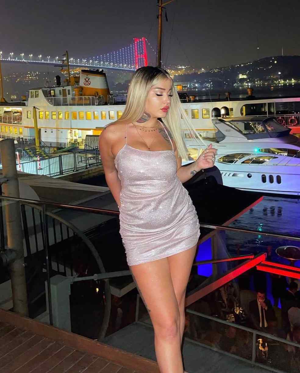 Woman in a light pink glittery mini dress posing on a marina promenade at night, with boats and a brightly lit bridge in the background.