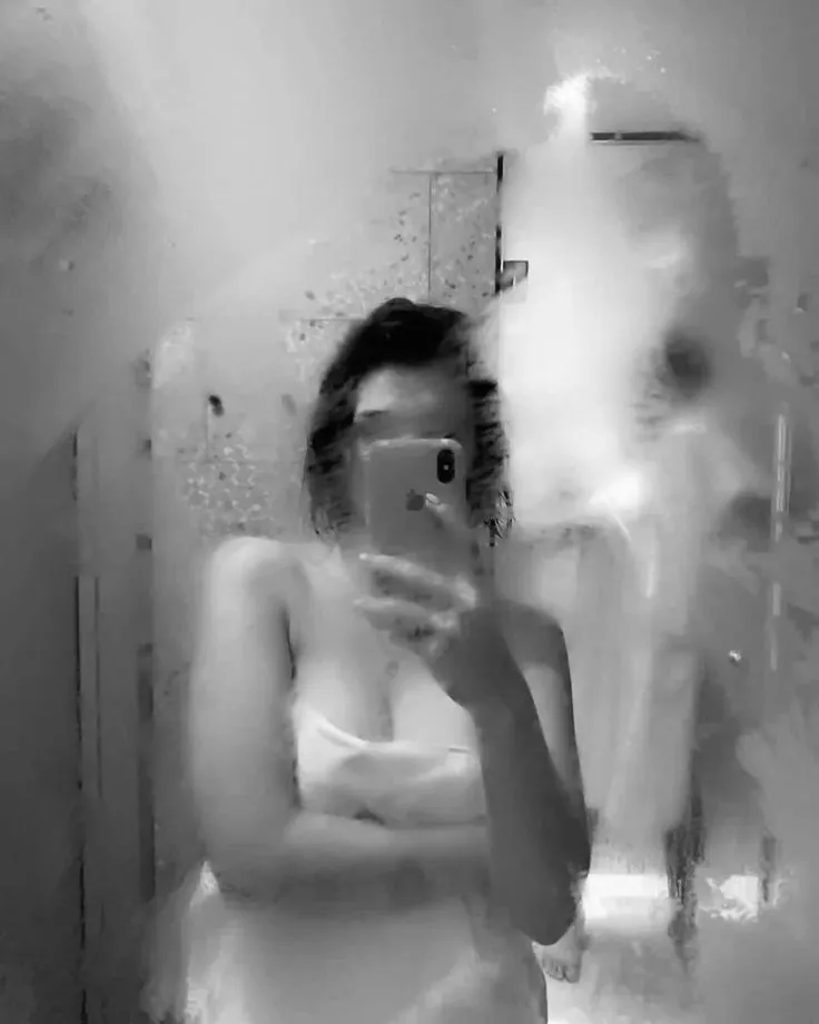 Person taking a mirror selfie with a smartphone in a dimly lit room; wearing a white tank top.