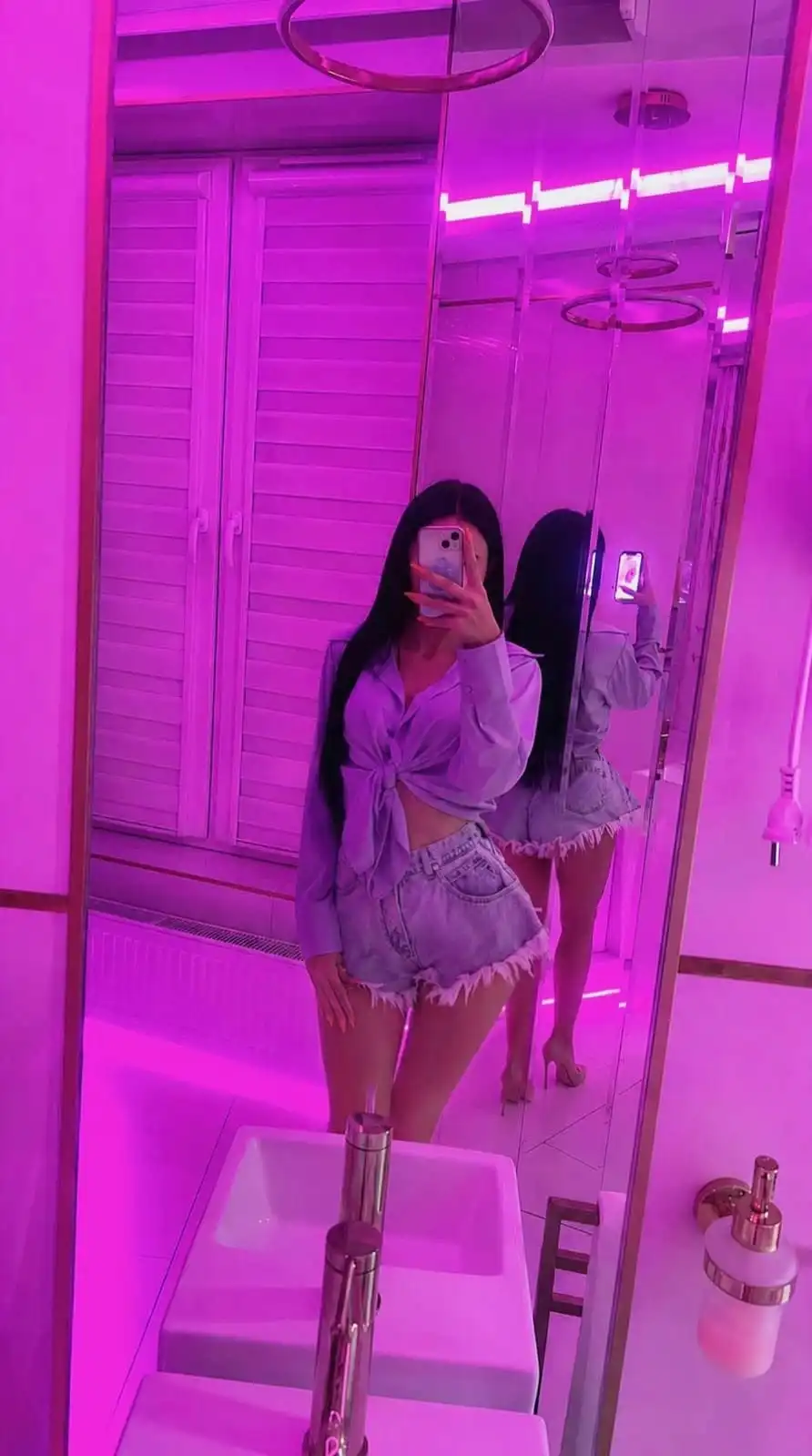 Person takes a mirror selfie in a pink-lit bathroom, wearing a tied lavender blouse and high-waisted frayed denim shorts, with a smartphone covering their face in front of a mirrored cabinet.