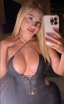 Self-portrait selfie of a blonde woman in a black lace plunging dress, taking a mirror photo with a pink iPhone.