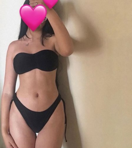 Person in a black strapless bikini taking a mirror selfie; face obscured by a pink heart overlay, standing against a beige wall.