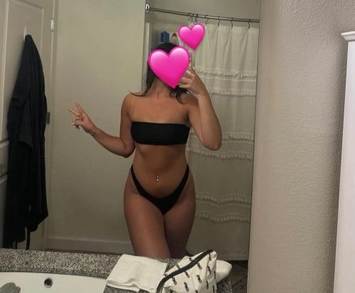 Person in a black bikini taking a mirror selfie in a bathroom; face obscured by pink heart stickers.