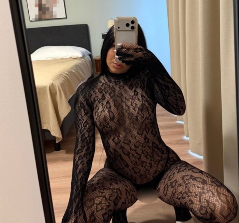 Person in a black lace bodysuit takes a mirror selfie with a phone, kneeling in a bedroom.