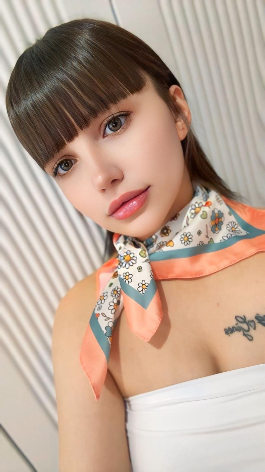 Young woman with straight bangs, wearing a floral scarf tied at the neck and a white strapless top.