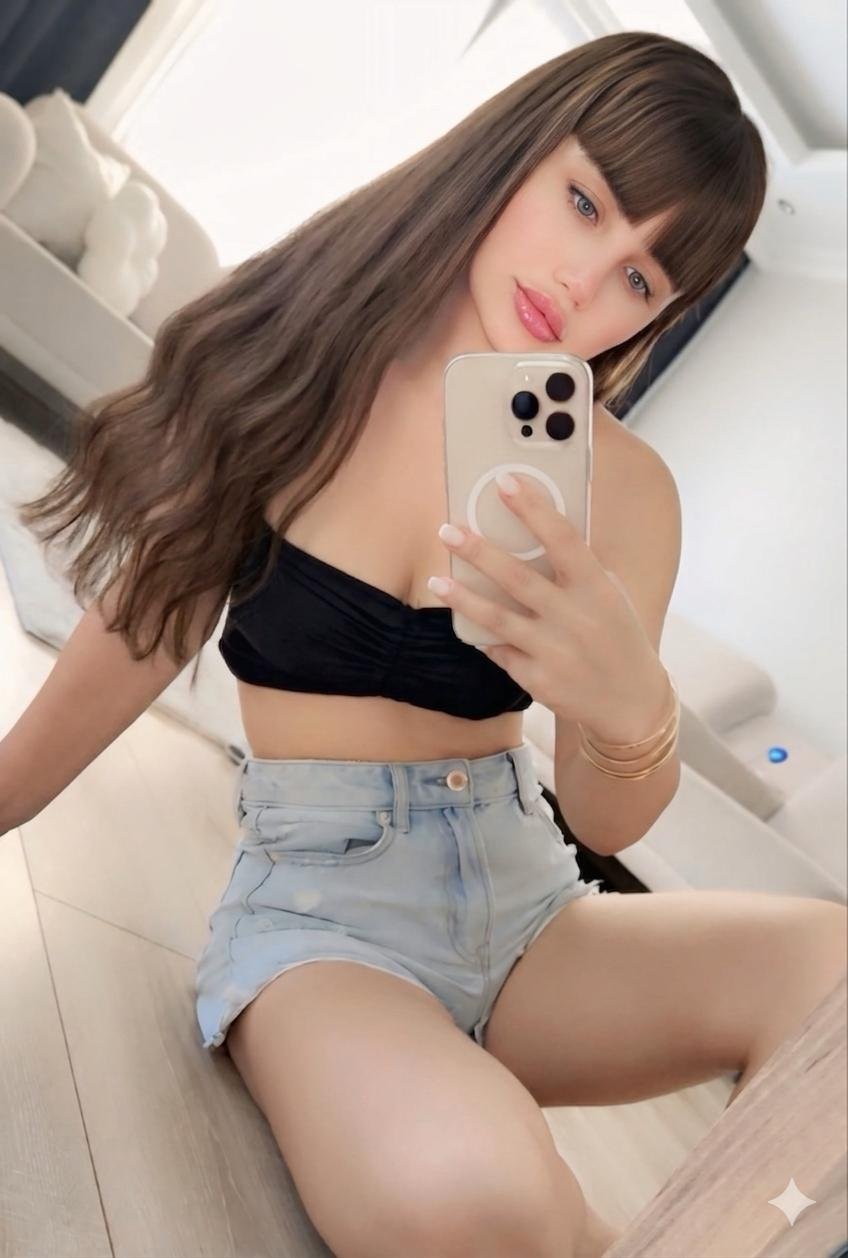 Woman taking a mirror selfie in a bright room, wearing a black crop top and light denim shorts, with long brown hair.