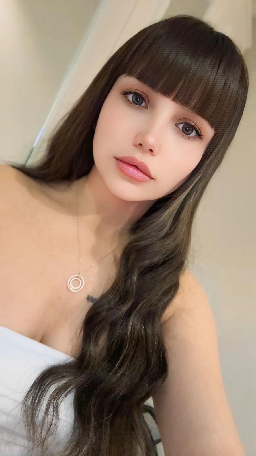 Young woman with long dark hair and bangs, wearing a white top and circular pendant necklace, looking at the camera.