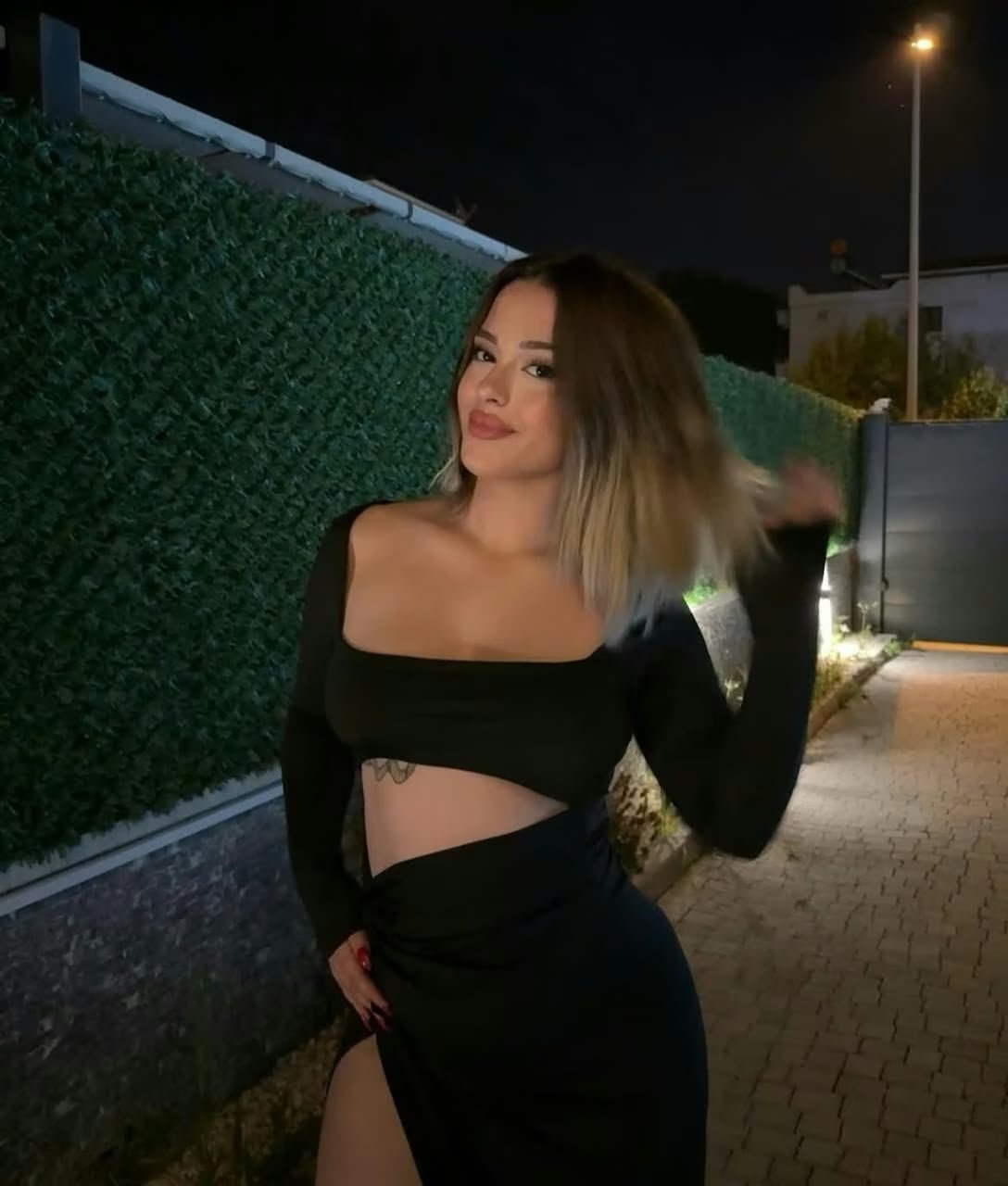 Woman in a black cropped top and high-slit skirt posing outdoors at night along a hedge-lined path, hand on hip, looking at camera.
