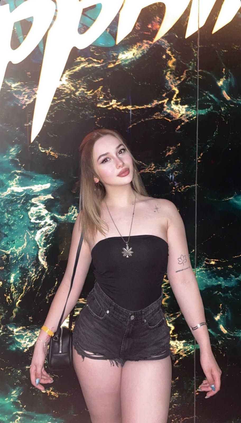 Young woman in a black strapless top and black denim shorts posing against a dark marble wall with white lettering; flower pendant necklace and crossbody bag, left arm tattoo visible.