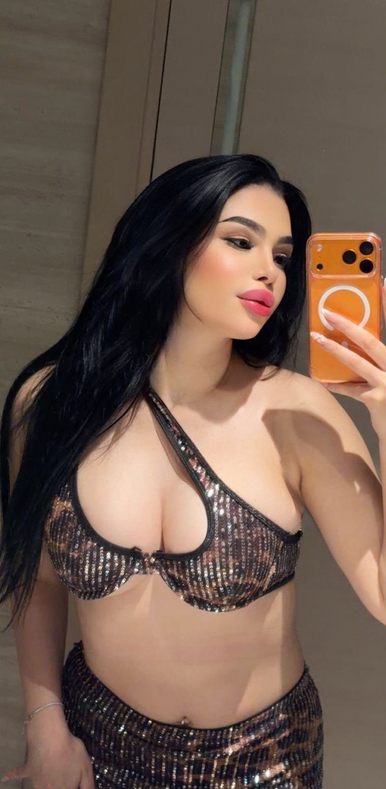 Woman taking a mirror selfie in a metallic sequined bikini top and matching skirt, holding an orange phone with a ring case.