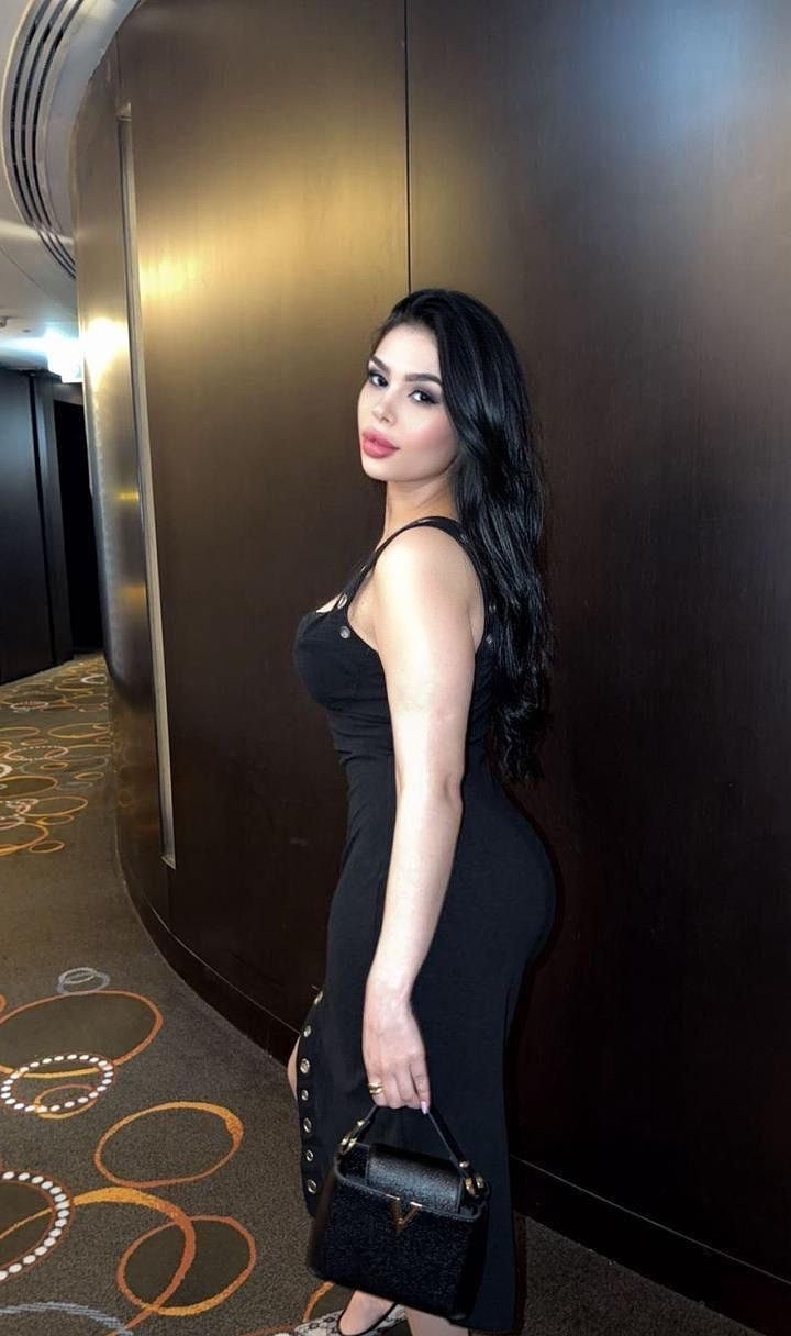 Woman in a black sleeveless dress stands sideways by a dark wall, holding a small black handbag with a gold accent.