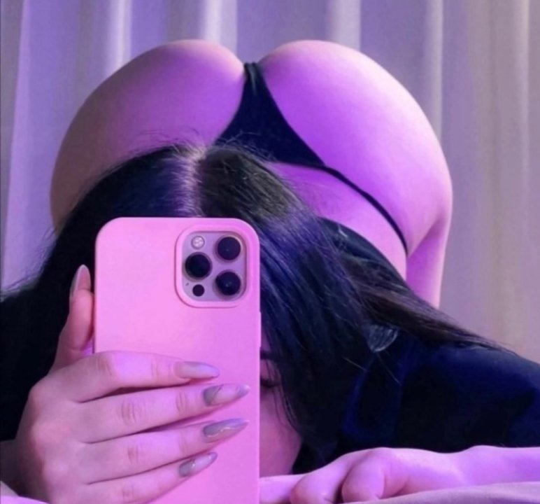 Person taking a mirror selfie with a pink phone, wearing a black bikini bottom under purple lighting, showing their backside.