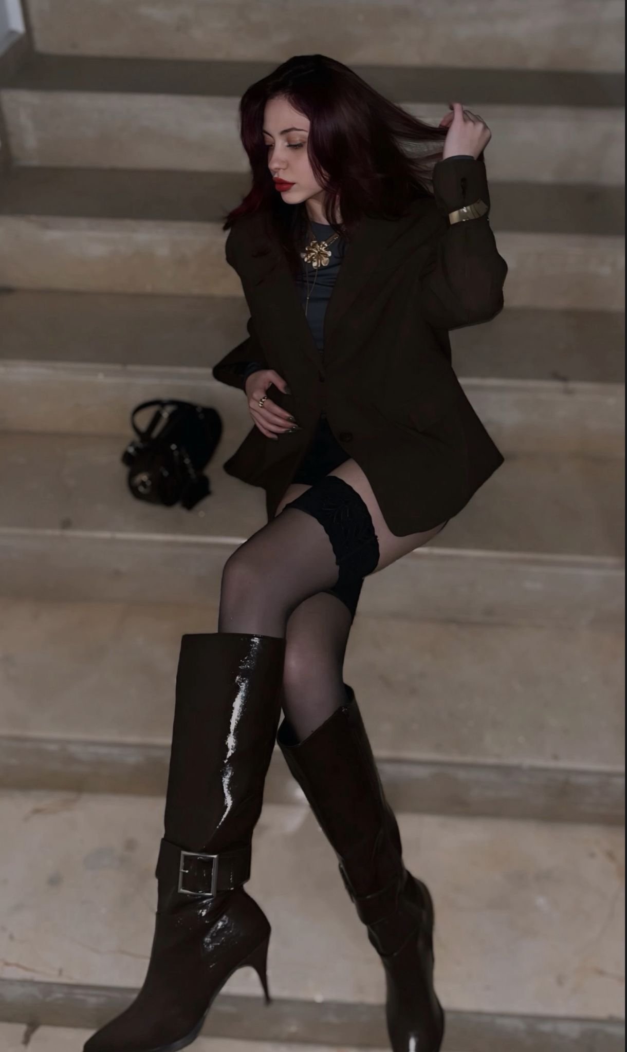 A woman with dark hair wearing a black blazer, blue top, thigh-high stockings, and tall boots posing on a concrete staircase indoors.