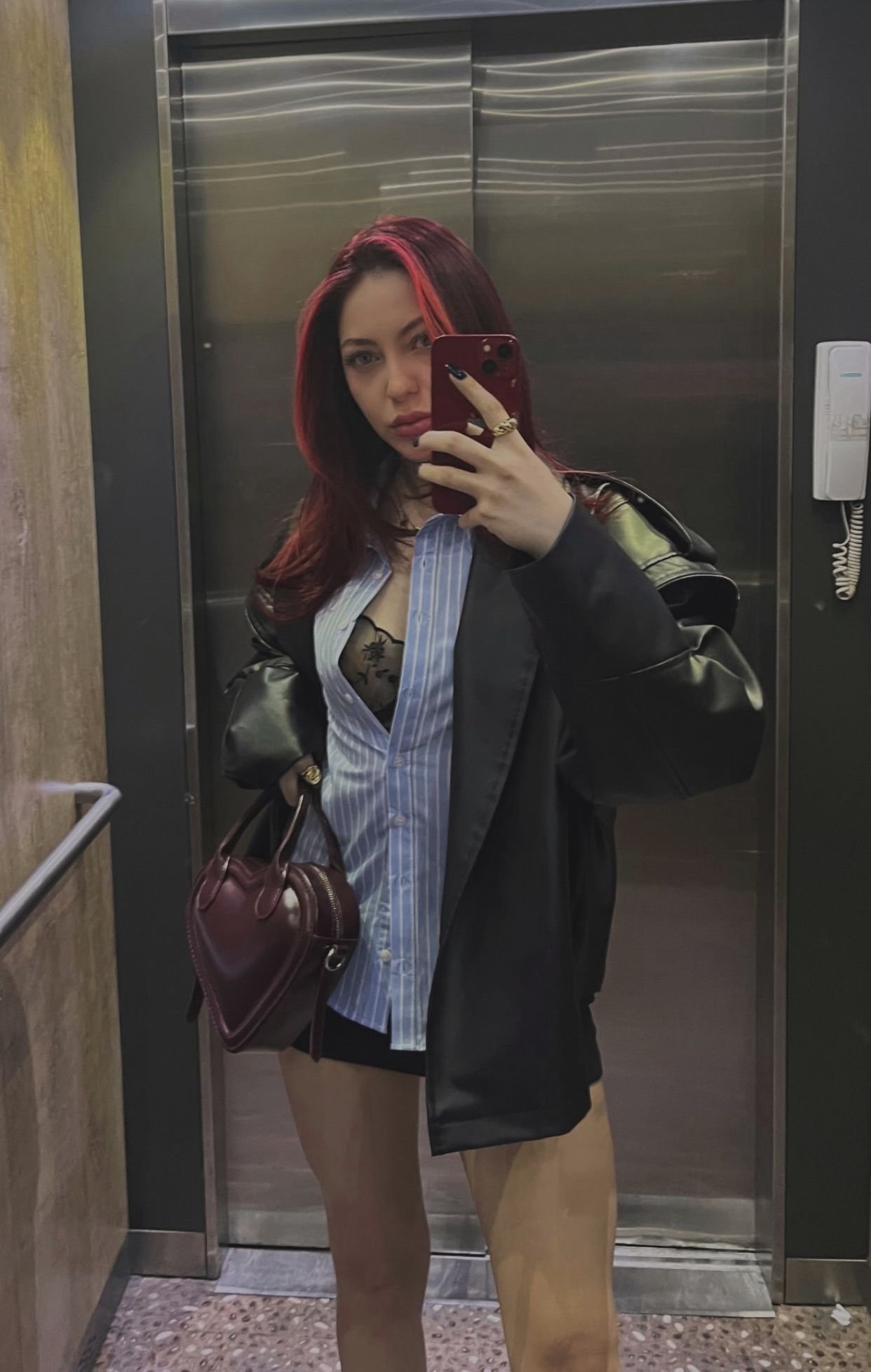 Young woman takes a mirror selfie in an elevator, wearing a blue striped shirt under a black coat, with red hair and a burgundy handbag visible at her side.