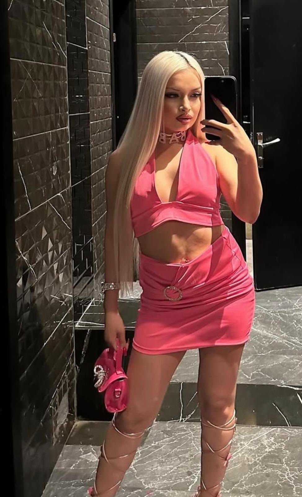 Woman with long platinum hair takes a mirror selfie in a marble-tiled bathroom, wearing a pink halter top and mini skirt, holding a pink purse.