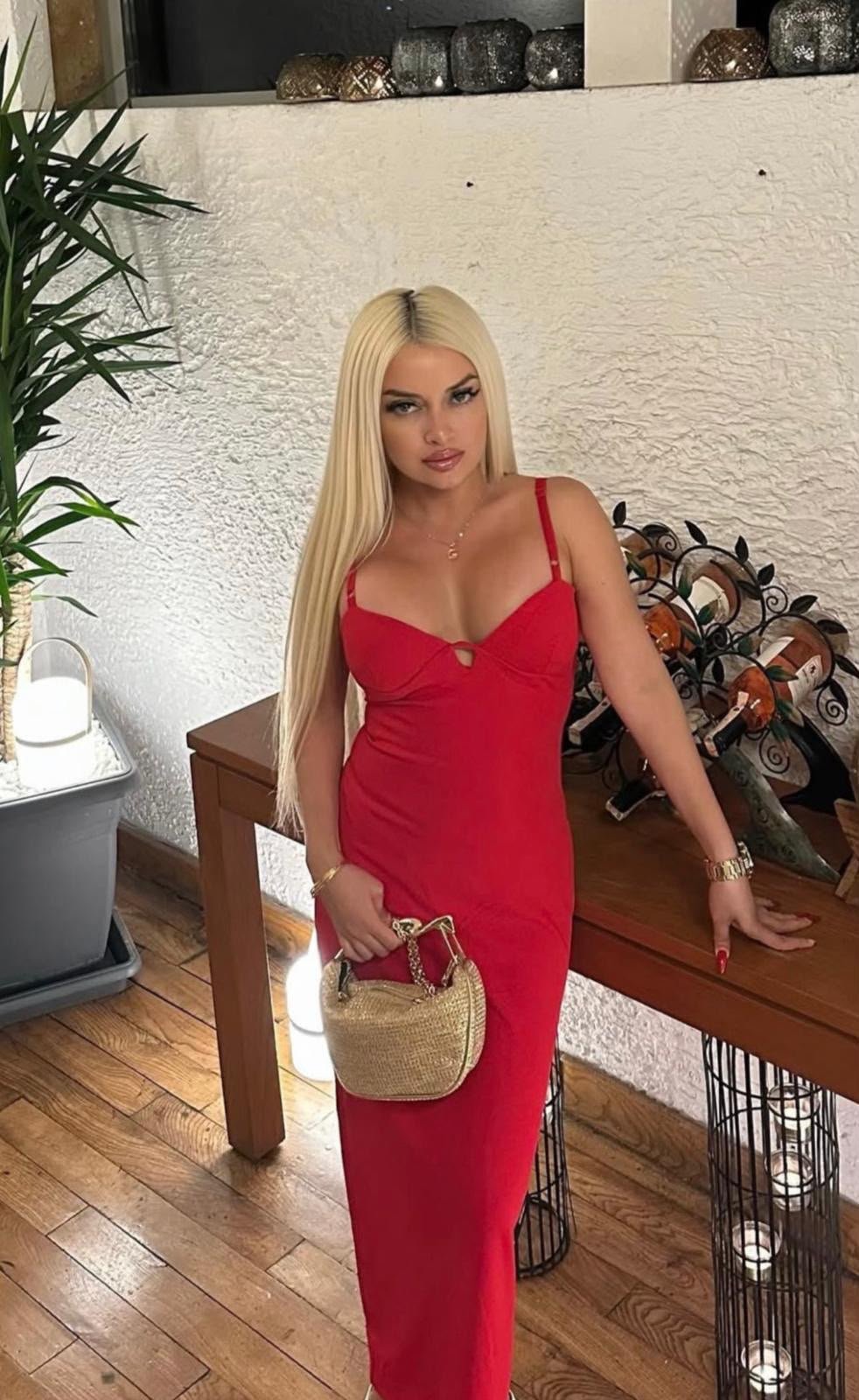 Woman in a red dress with long blonde hair posing indoors, holding a straw handbag near a wooden table.