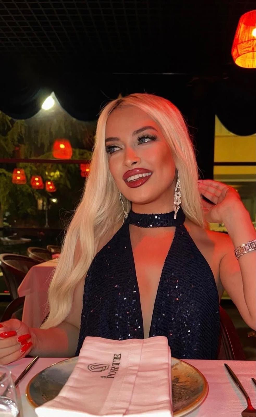Woman with long blonde hair and a sparkly black halter dress sits at a restaurant table, smiling warmly under warm lighting.