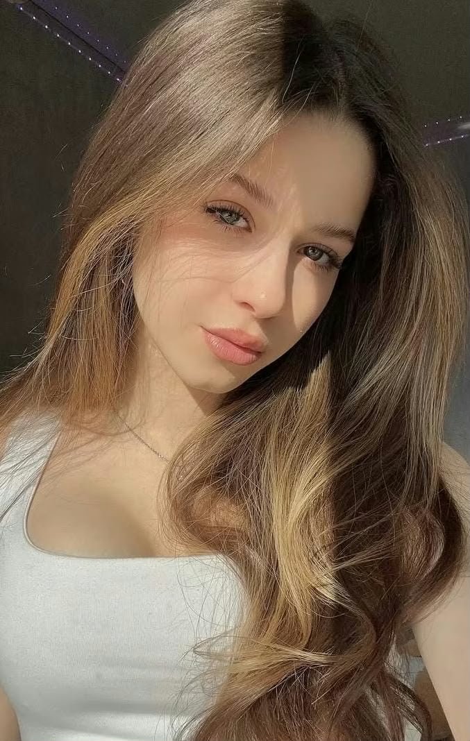 Close-up of a young woman with long light brown hair wearing a white top, bathed in warm sunlight.