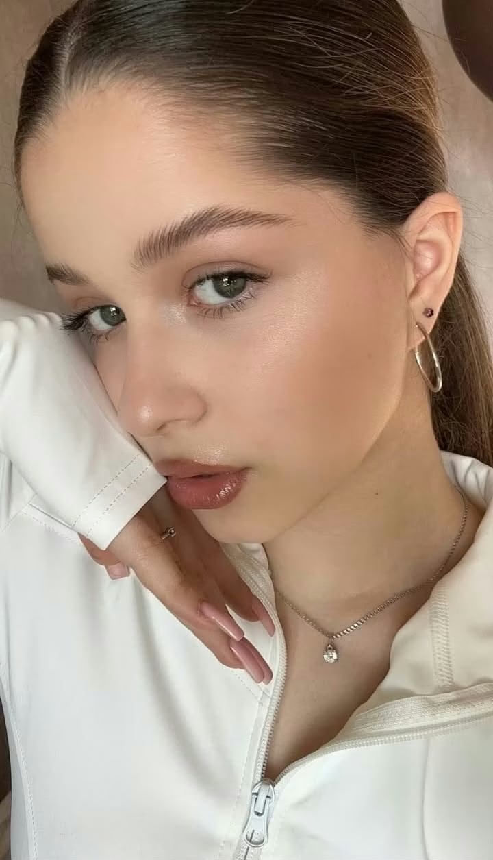 Close-up portrait of a young woman with slicked-back hair, subtle makeup, wearing a hoop earring and a pendant necklace.