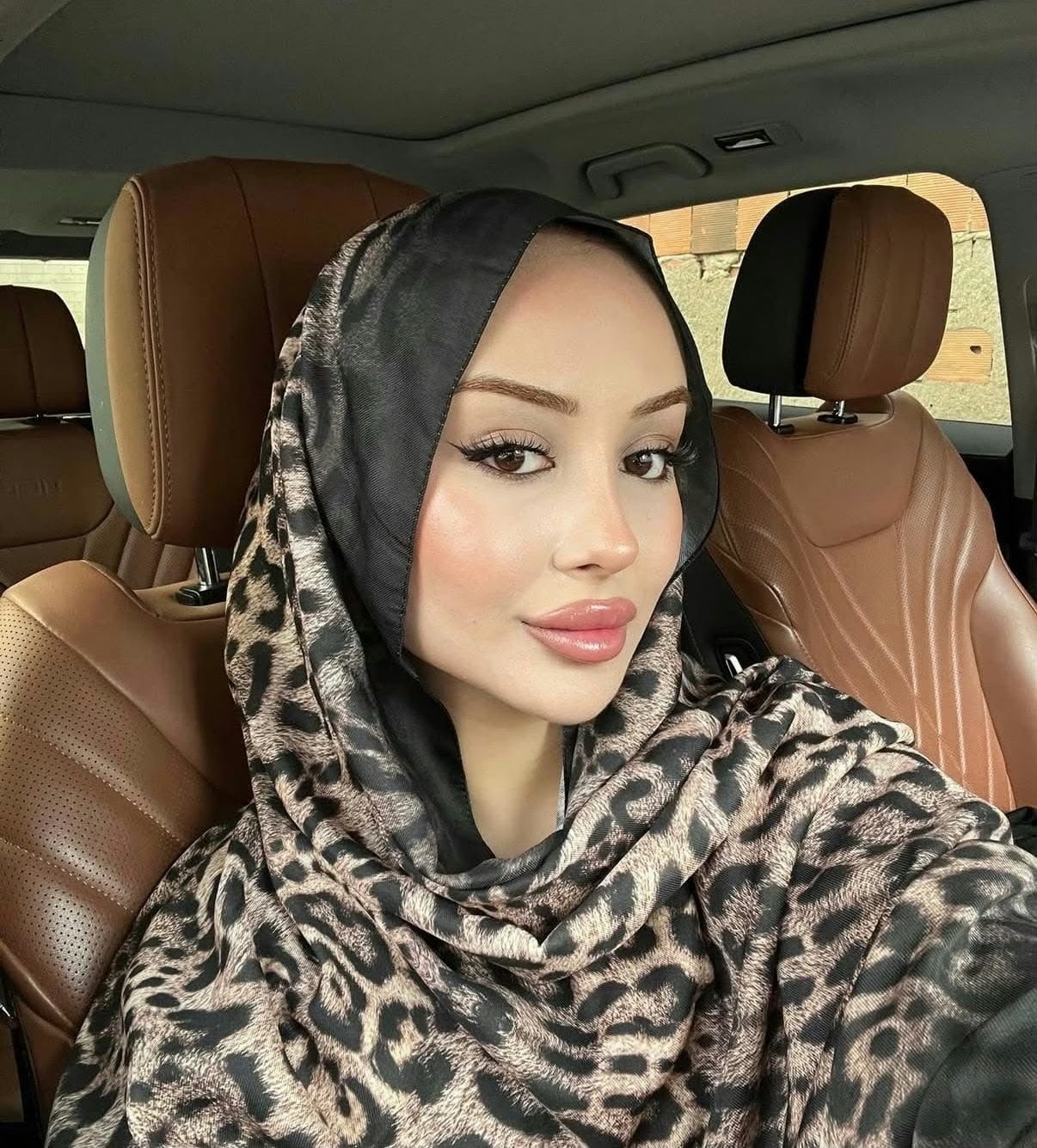 Woman wearing a leopard-print hijab and makeup sits in the front seat of a car with brown leather seats behind her.
