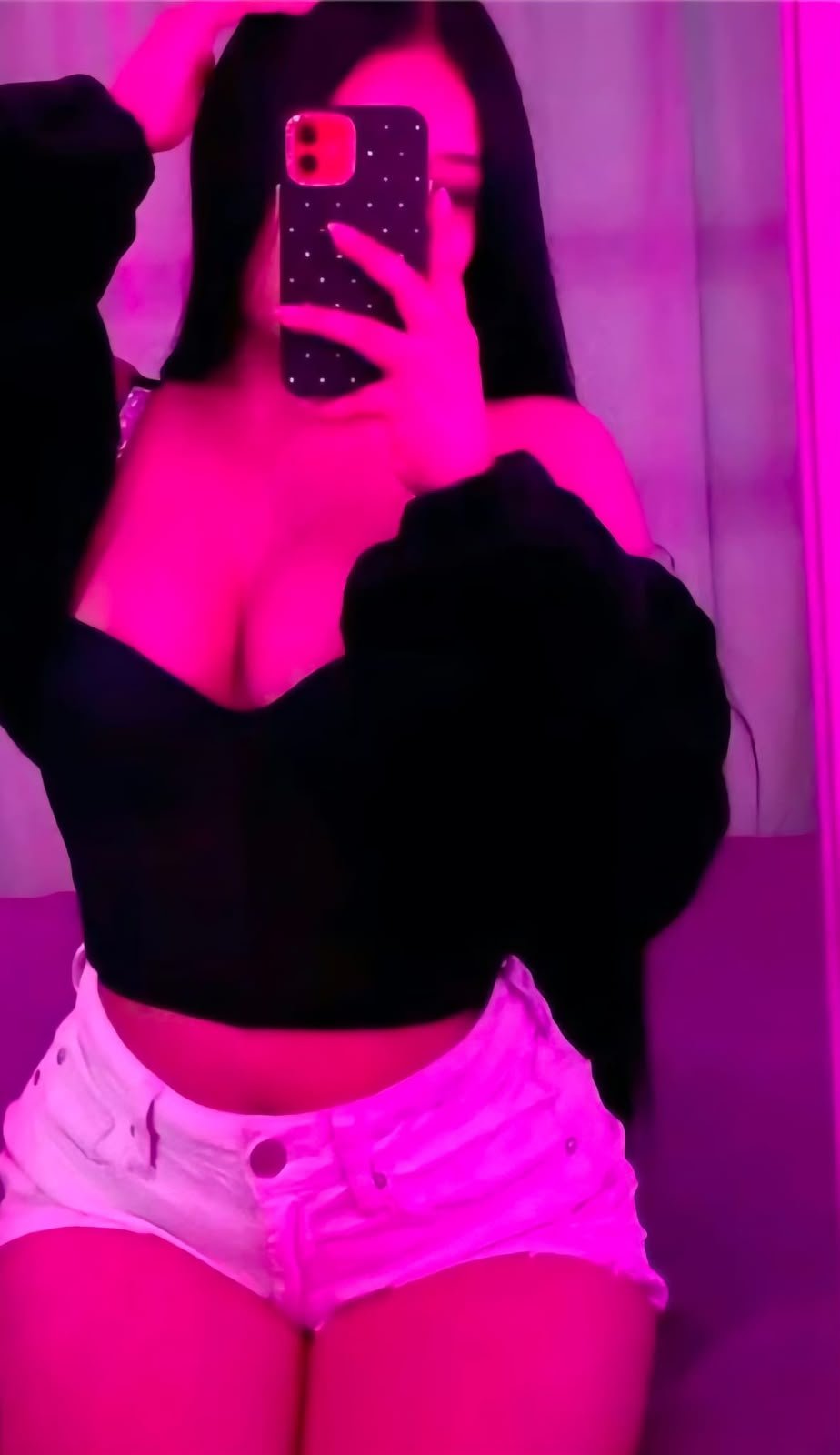 Person taking a mirror selfie in a pink-lit room, wearing a black off-the-shoulder top and white shorts, holding a phone with a dark star-dotted case.