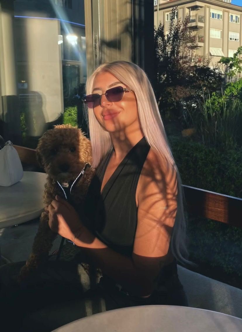 Woman with long blonde hair wearing sunglasses sits at an outdoor table, holding a small brown poodle.