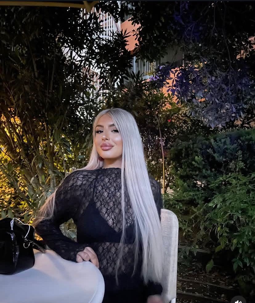 Woman with long silver hair in a black lace top sits at a table outdoors at night, surrounded by greenery.