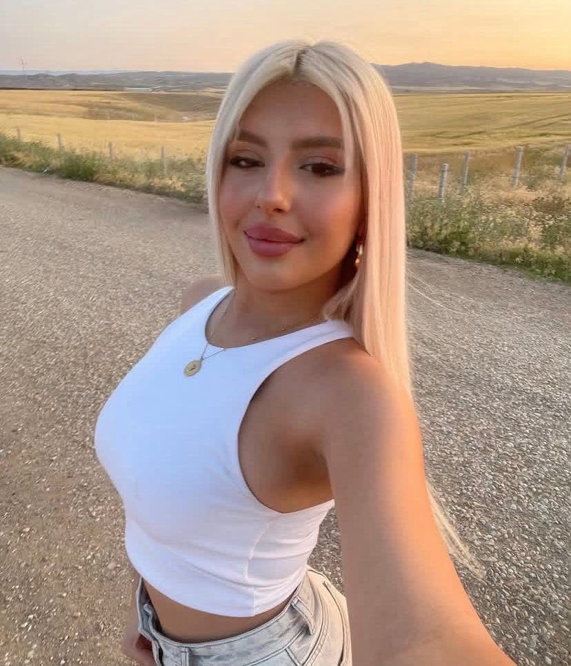 Young woman with long blonde hair taking a selfie on a rural road at sunset, wearing a white tank top.