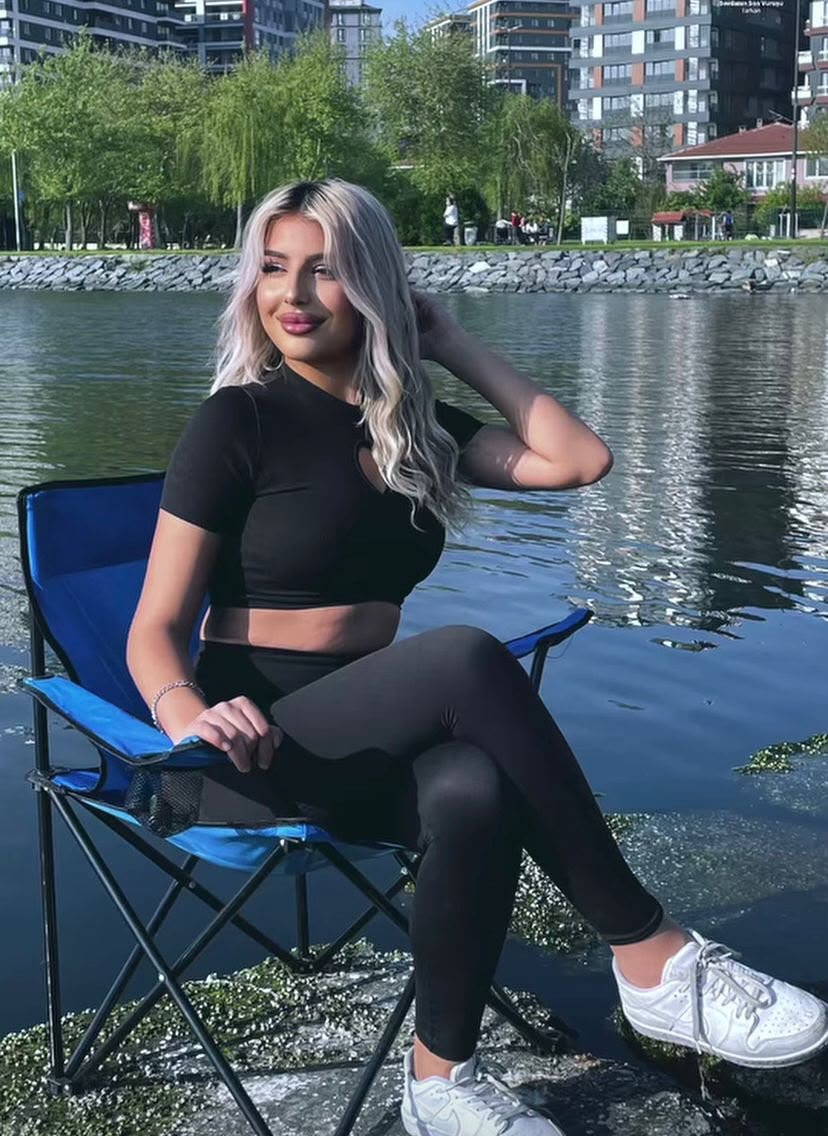 Woman with long blonde hair sits on a blue folding chair by a lake, city buildings visible in the background.