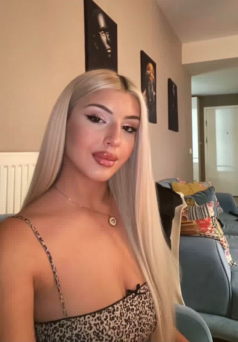 Woman posing for a selfie in a home setting, with long platinum blonde hair and a leopard-print top.