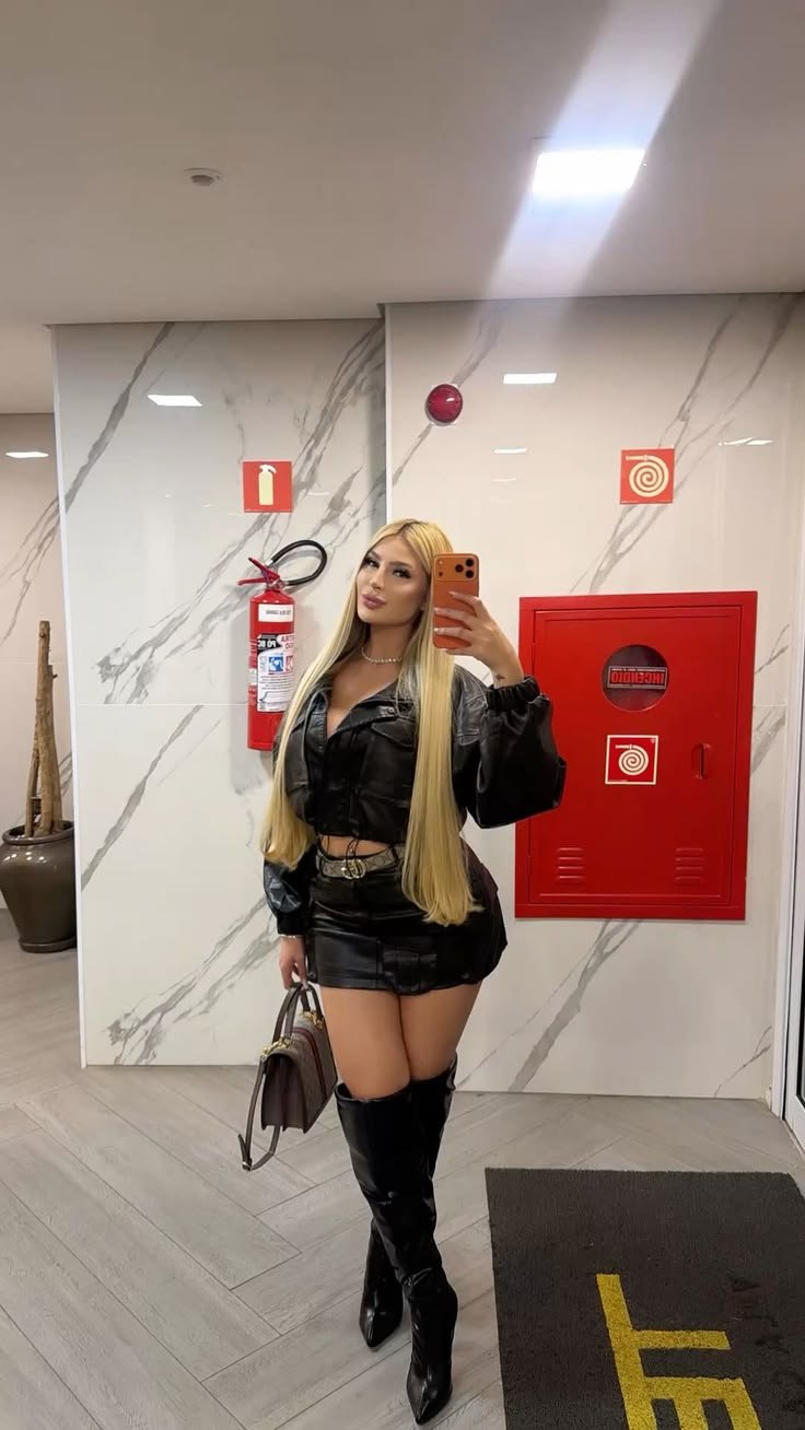 Woman in a black leather outfit with thigh-high boots taking a full-body mirror selfie in a marble-walled hallway, holding a handbag.