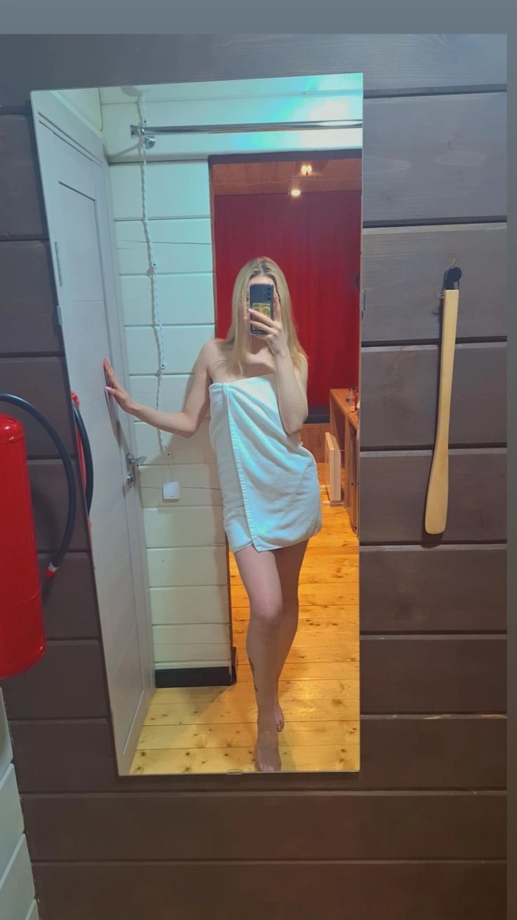 Woman in a white towel taking a full‑length mirror selfie with her phone, barefoot in a wood-paneled room.