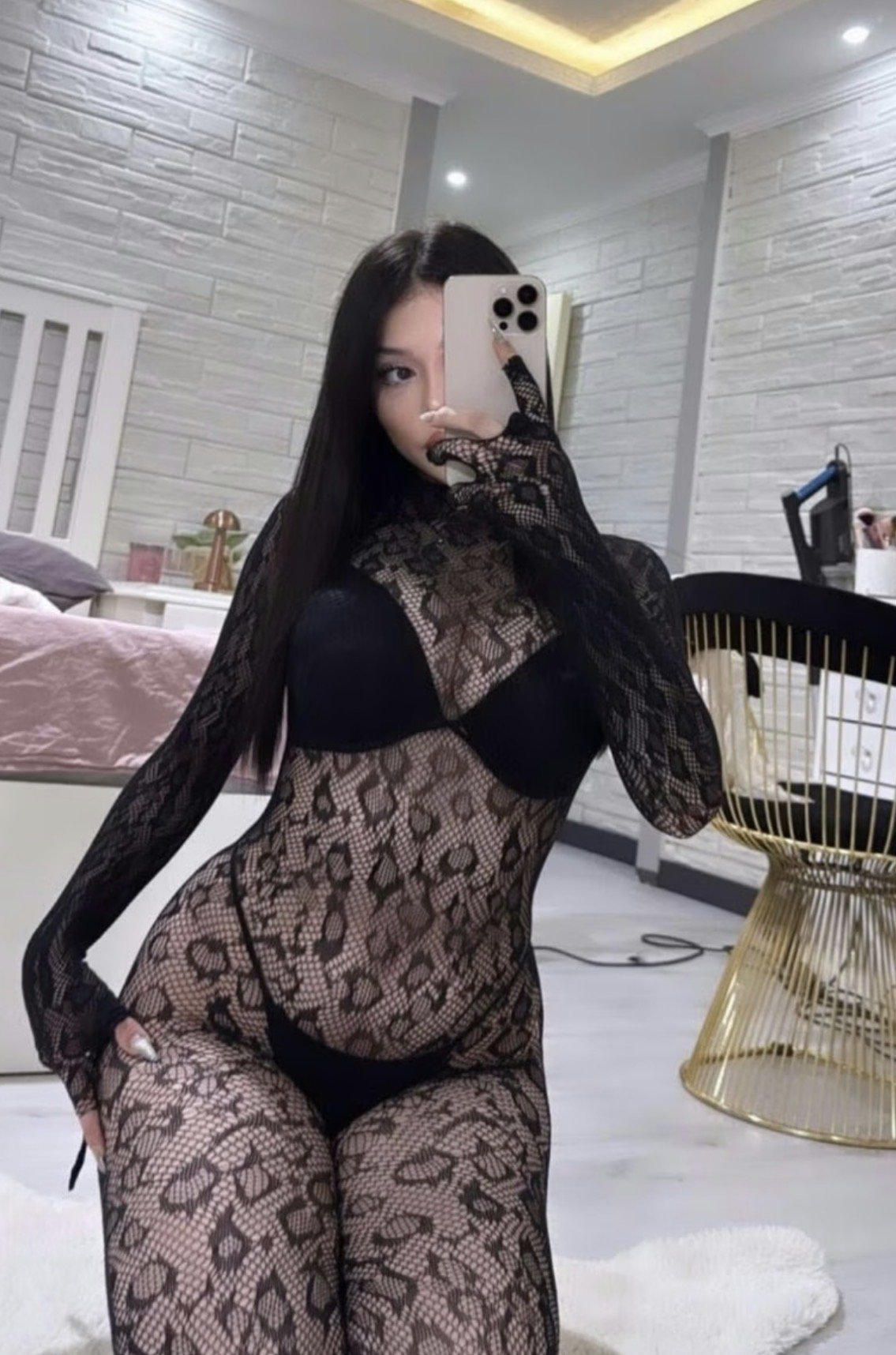 Woman in a black lace bodysuit taking a mirror selfie in a bedroom, phone covering part of her face.