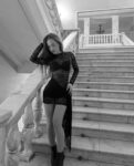 Woman in a black lace dress posing on a grand marble staircase, her hand in her hair and gaze downward.