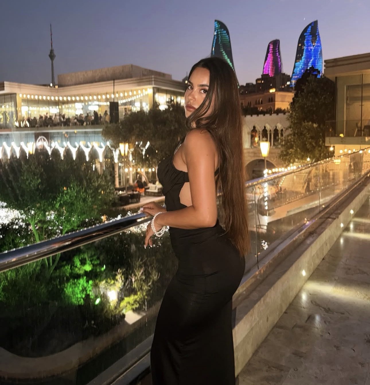 Woman in a black evening dress posing at a railing on a night-lit terrace, with a cityscape and colorful lit towers behind her.