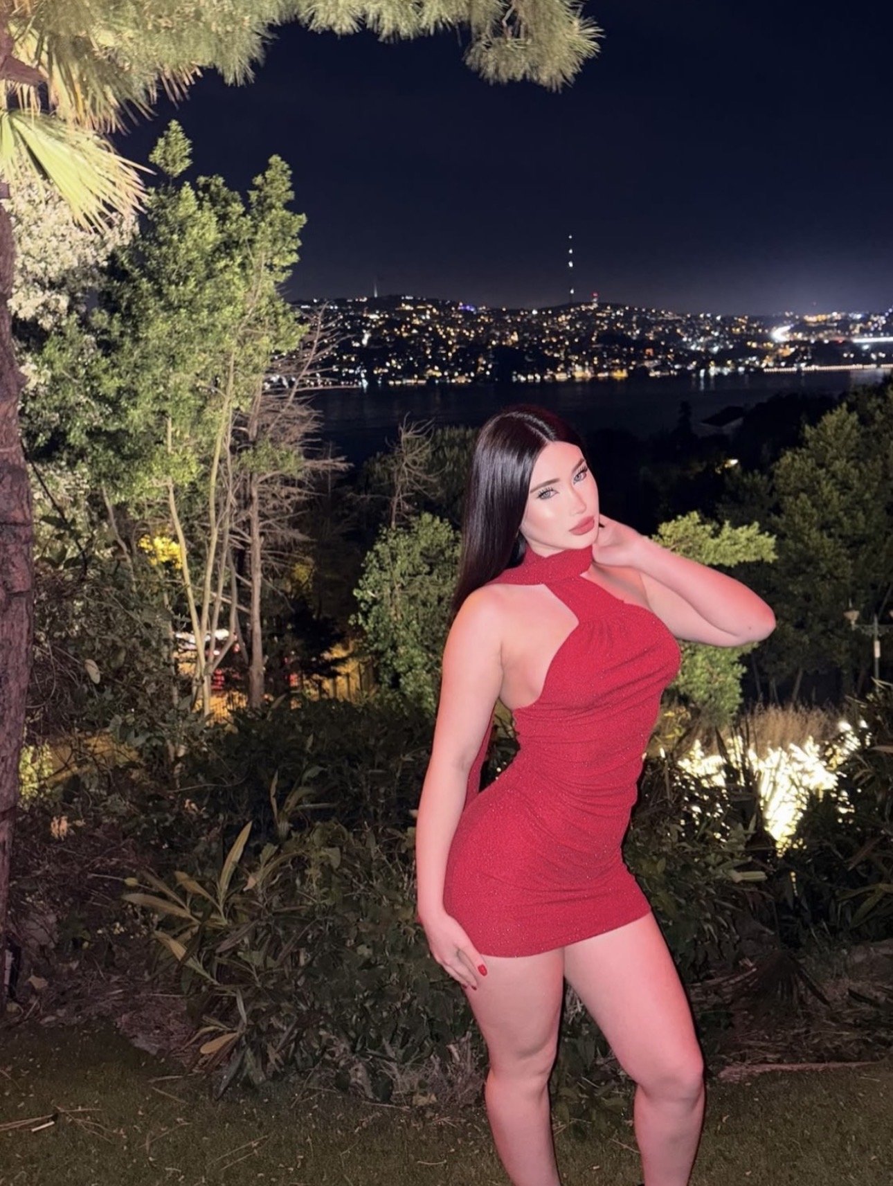 Woman in a red halter dress posing outdoors at night with city lights in the background and trees surrounding her