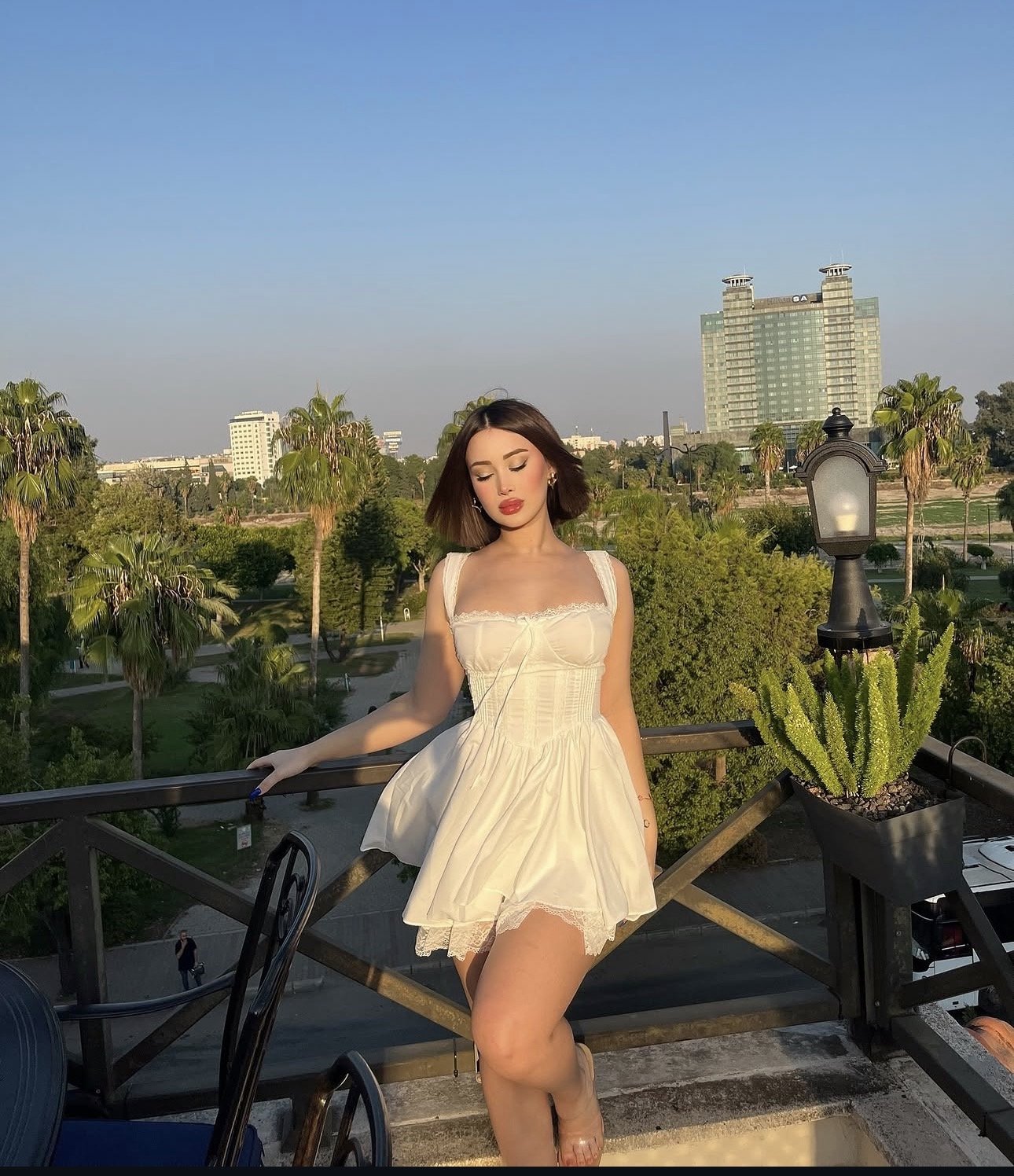 Woman in a white dress posing on a balcony with a park and palm trees in the background. Sunset light highlights her makeup and posture.